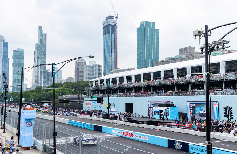 Racecars are lined up along a stretch of road in front of grandstands.