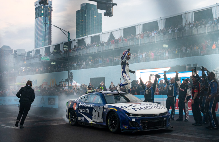 A NASCAR driver wearing a fire suit and helmet stands on the roof of his car with his fist in the air. 